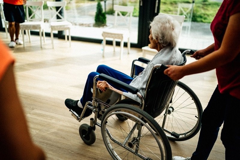 An older person seated in a wheelchair being assisted by a carer